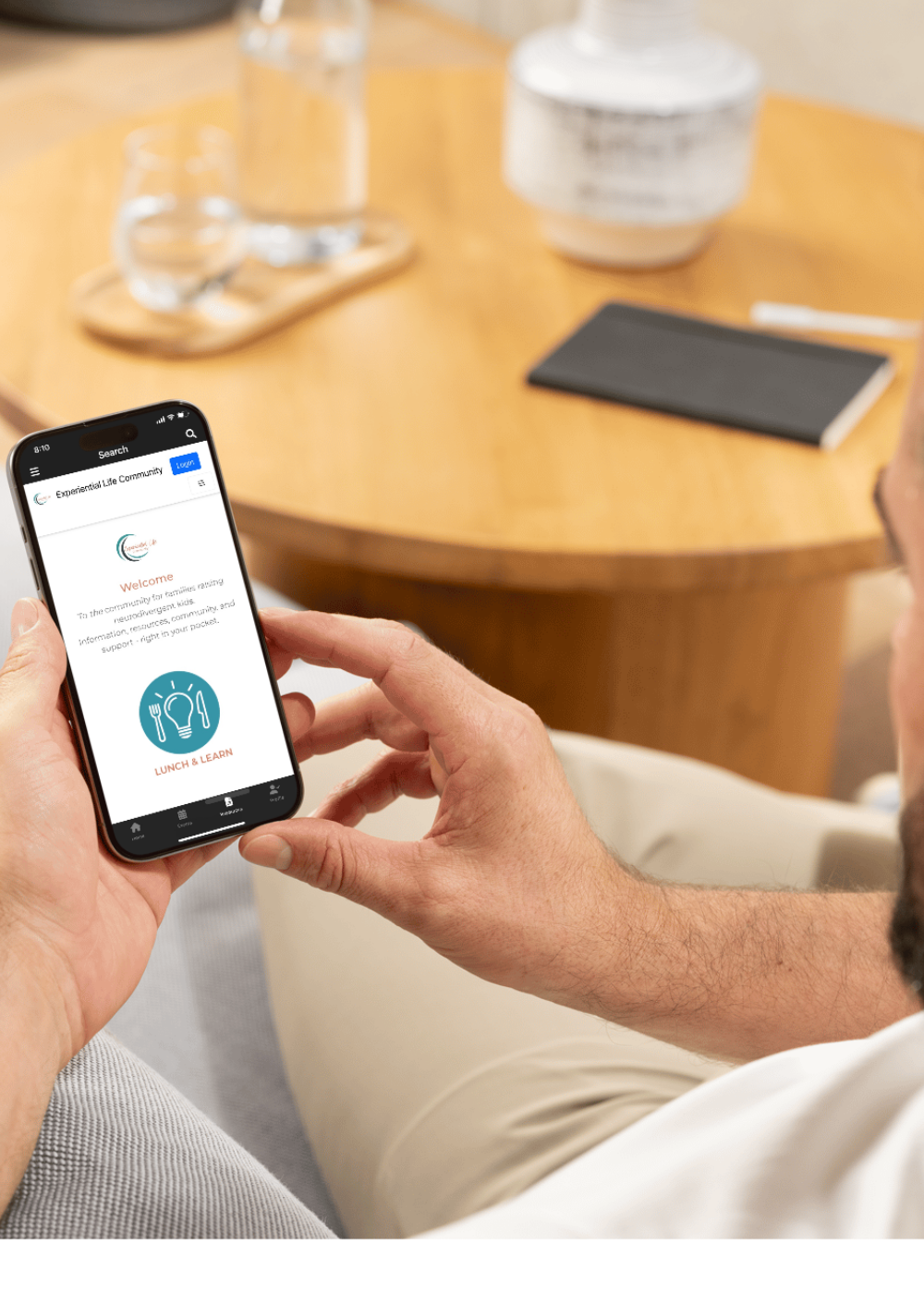 A white man sits on the couch with a coffee table to his right. In his hand is his cell phone with the Experiential Life App dashboard on the screen.