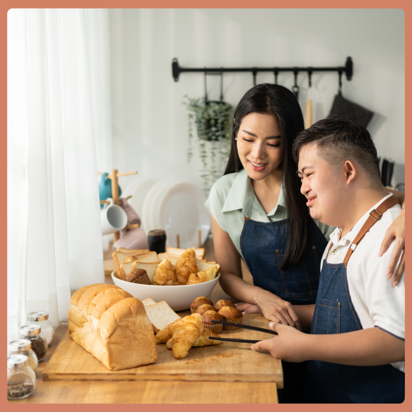 An Asian man with Down syndrome stands in front of a counter with freshly baked bread, holding a knife. His sister has one arm around his shoulder and the other hand is guiding his to cut the bread safely.