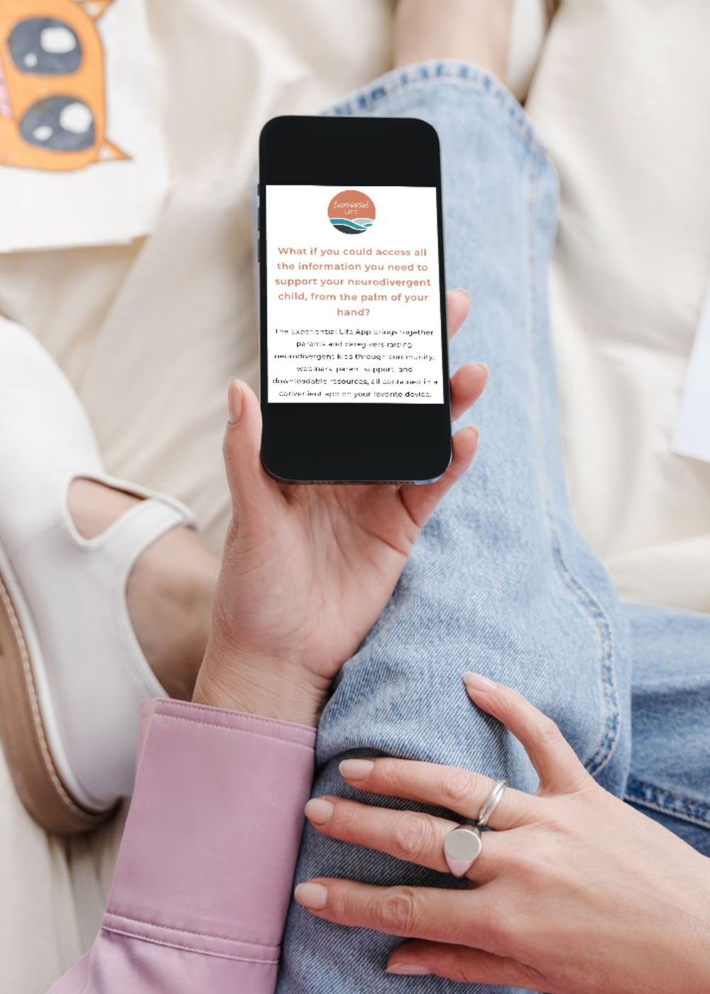 A woman sits on a bed with a white comforter beneath her. She is holding her iPhone looking at the Experiential Life website.