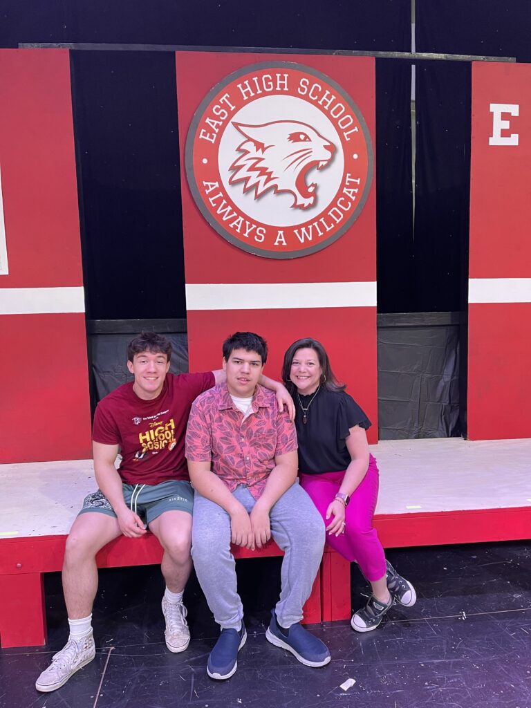 My middle son (we call him O around here) sits on the left wearing his High School Musical t-shirt and shorts. C, my autistic son sits in the middle, wearing a Hawaiian shirt and grey pants. I sit on the right, with a black blouse and hot pink pants. We are sitting on the stage, with the East High School logo and mascot behind us - the same one from High School Musical.