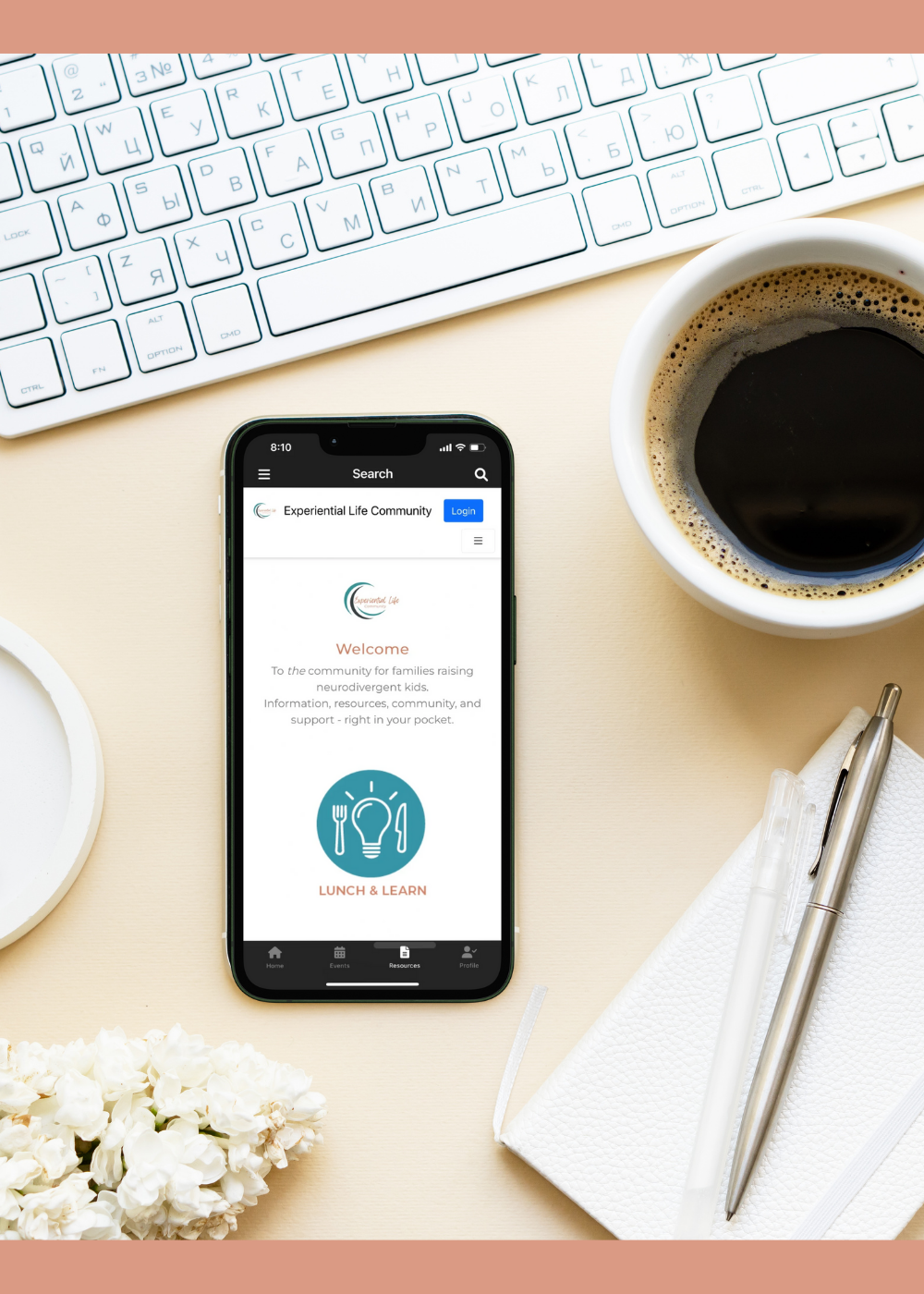 A cell phone sits on a desk just under a white keyboard with a cup of coffee and a notebook and pen sitting to the right of it. On the cell phone screen is the Experiential Life App dashboard.
