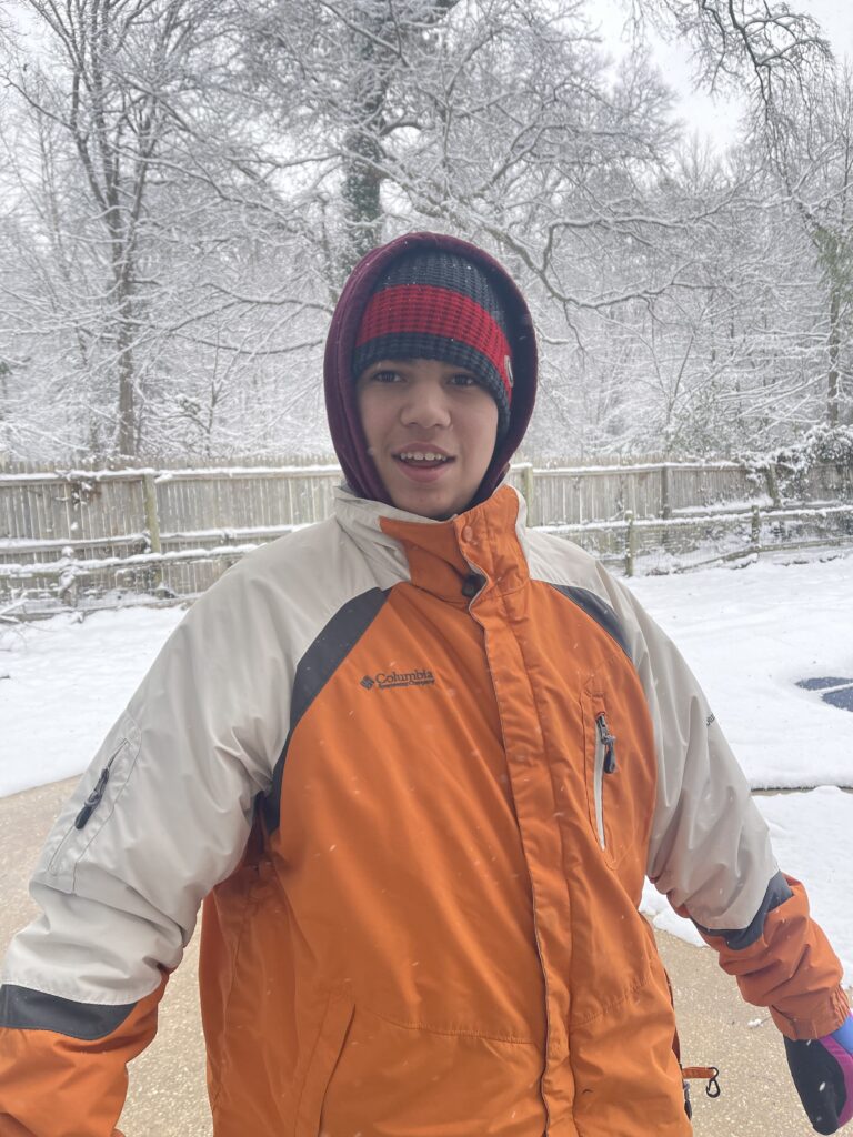 C is a white teenaged boy standing outside in the middle of a snow storm. He is wearing a winter hat and an orange and beige snow coat. There are trees covered in snow behind him.
