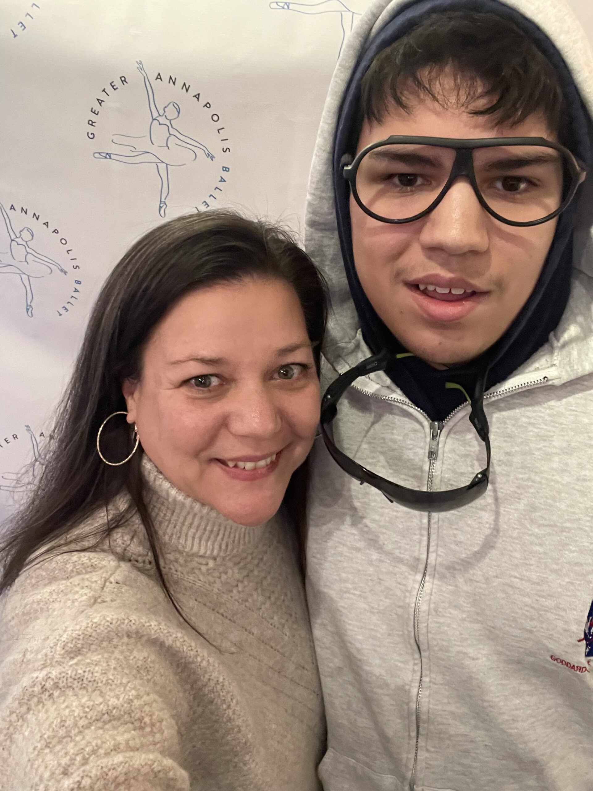 Christy is a white woman wearing a beige sweater and her dark hair tucked behind her ears. She is standing to the left of her autistic teen, who is white, much taller, and wearing a grey zip hoodie. His hood is covering almost all of his dark brown hair and he is wearing black framed glasses. His sunglasses hang on a lanyard over his hoodie.